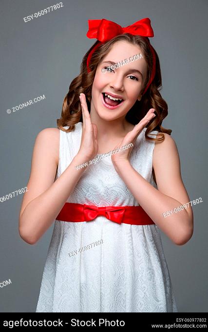 Beautiful teenage girl with long curly hair and red ribbon bow on head wearing white dress. Happy expression. Studio portrait on grey background