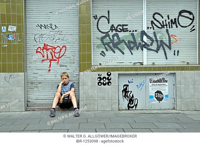 Lonely nine-year-old boy in front of a closed shop smeared with graffiti, Germany, Europe