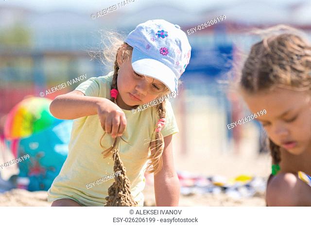 Four-year girl builds a sand castle on the beach of the seaside, beside the other girl sitting