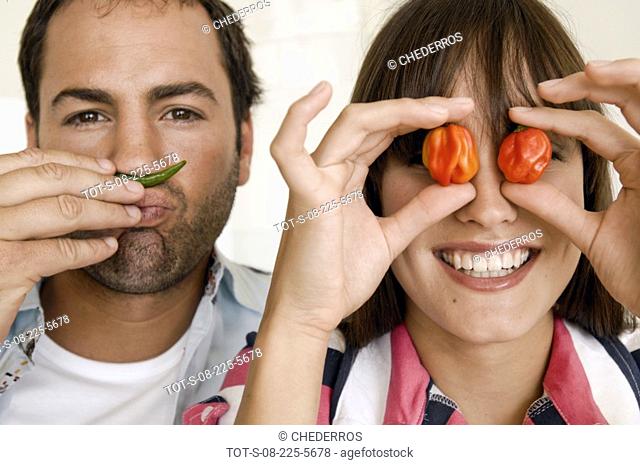 Close-up of a young woman covering her eyes with red chili peppers with a young man making mustache with a green chili pepper