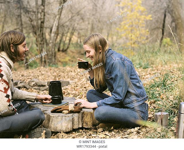 Smiling couple sitting on the ground in a forest, drinking coffee