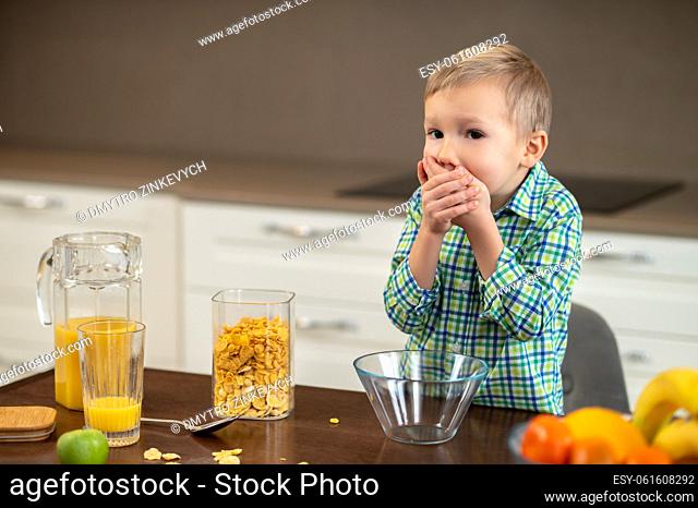 Waist-up portrait of a kid standing at the table and closing his mouth with both hands