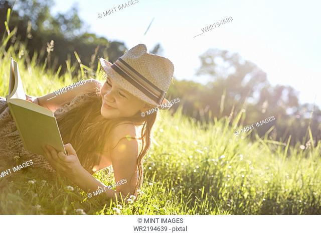 A young girl in a straw hat lying on the grass reading a book