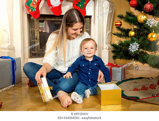 Cute baby boy with his mother opening gift boxes under Christmas tree at living room