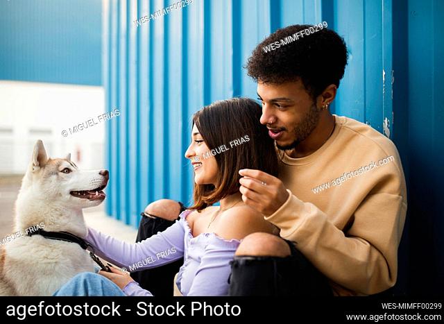 Smiling couple sitting with dog while leaning on blue wall