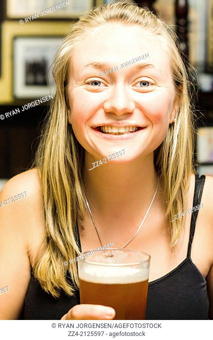 Lifestyle photo of a smiling young attractive blond woman in mid 20s holding and drinking a pint of amber ale beer inside British bar