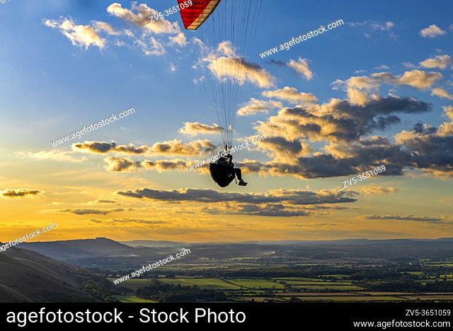 Paraglider flying against the setting sun & scattered clouds, above the South Downs National Park. Brighton, East Sussex. Uk