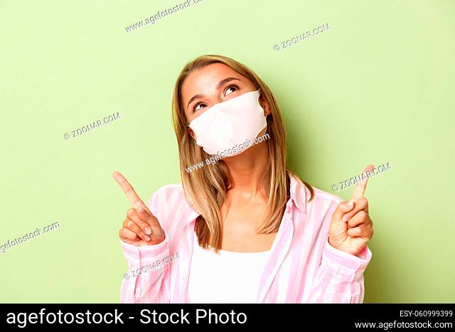 Concept of covid-19, social-distancing and lifestyle. Close-up of beautiful dreamy girl in face mask, looking and pointing up, standing over green background