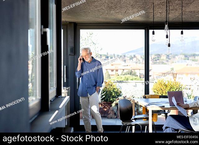 Senior man talking on mobile phone while standing with hands in pockets at home