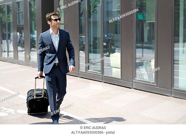 Businessman with sunglasses pulling luggage, walking in city