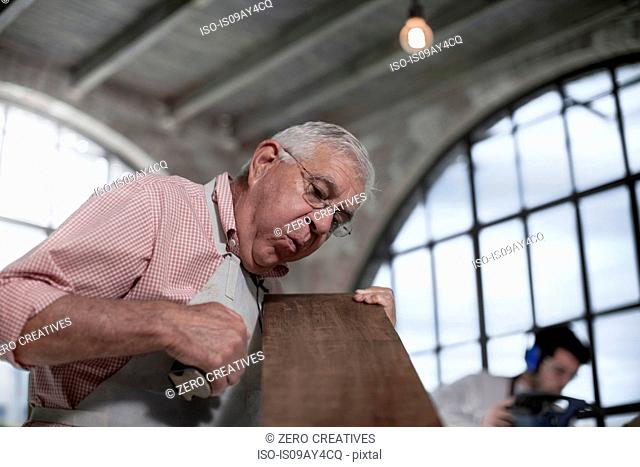 Senior carpenter blowing sawdust from wood plank in workshop
