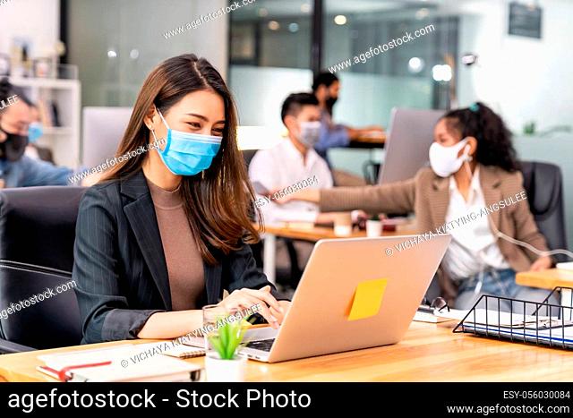 Portrait of asian office employee businesswoman wear protective face mask work in new normal office with interracial team in background as social distance...