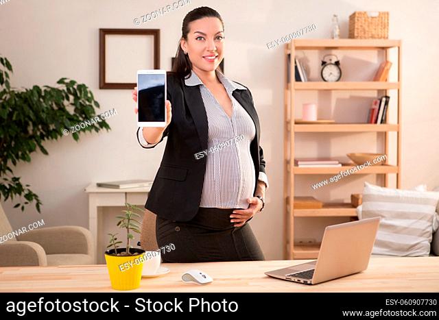 Pregnant business woman showing smart or mobile phone to camera while working at home. Excited lady showing phone with blank screen for your ideas and desires