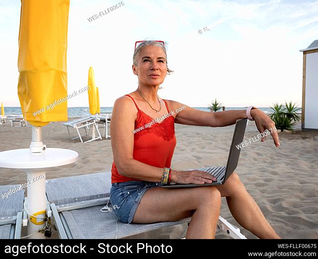 Thoughtful senior businesswoman sitting with laptop at beach