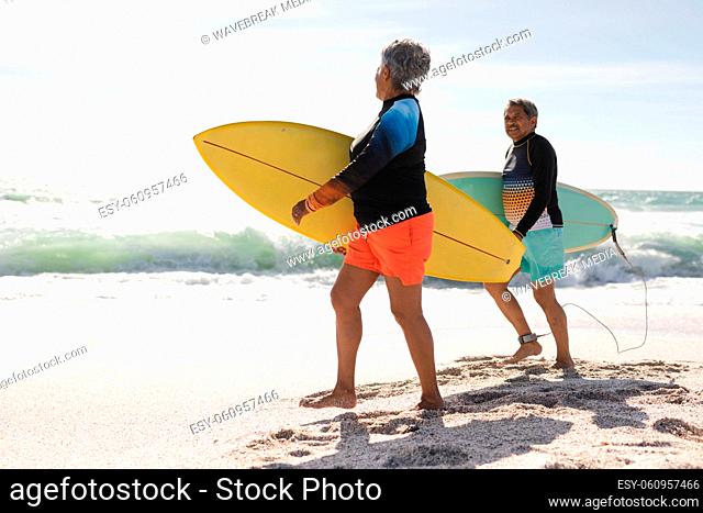 Full length side view of multiracial senior couple walking with surfboards at beach during sunny day
