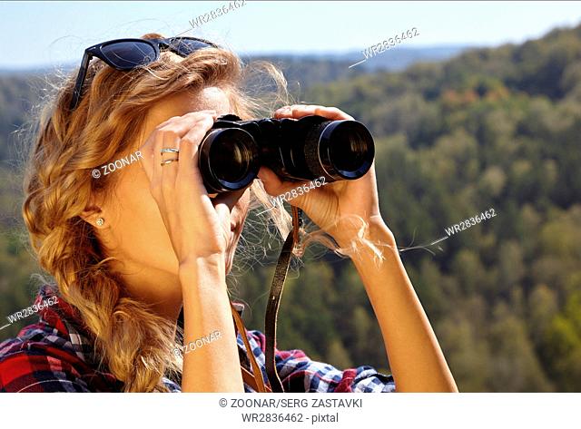 Young blonde woman tourist  on a cliff looking through binoculars on the autumn landscape