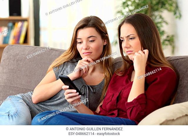 Two upset friends viewing on line content in a smart phone sitting on a sofa in the living room at home