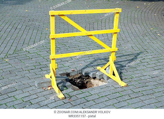 A homemade wooden yellow barrier stands over a pit in an urban granite pavement