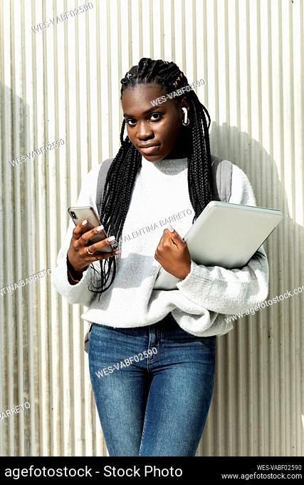 Portrait of a young woman standing at a wall with cell phone and laptop