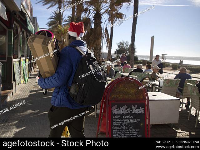 11 December 2023, Spain, Palma: A sunglasses salesman in a Santa hat walks past a restaurant on Arenal beach. Mallorca recorded a record temperature of 24...