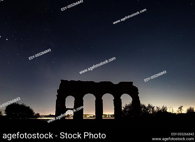 The ruins of an ancient Roman aqueduct at night with starry sky. In the background, the wake of a train running fast on the tracks