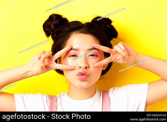 Carefree asian girl showing v-signs and kissing lips, standing on yellow background
