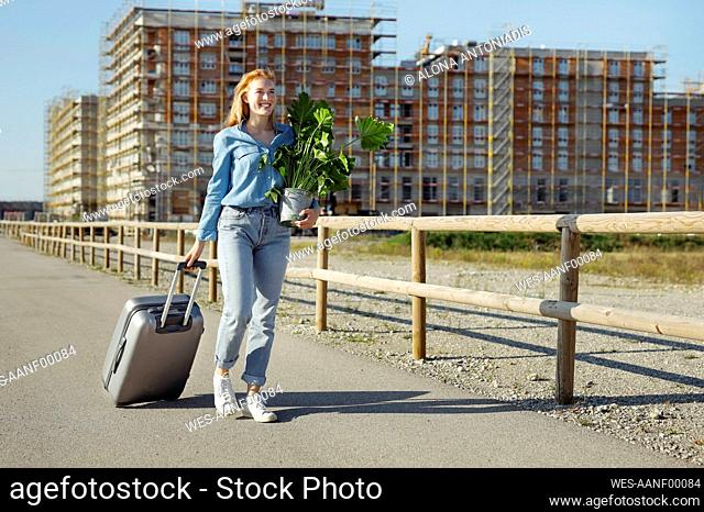 Young woman walking with plant and wheeled luggage on sunny day