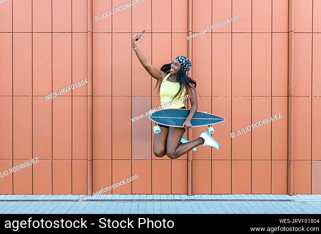 Smiling woman with skateboard jumping while taking selfie through smart phone in front of wall