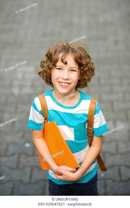 The fair-haired curly schoolboy looks in the camera and smiles. The boy has a nice face, a fair hair, an open look. It has behind shoulders a satchel