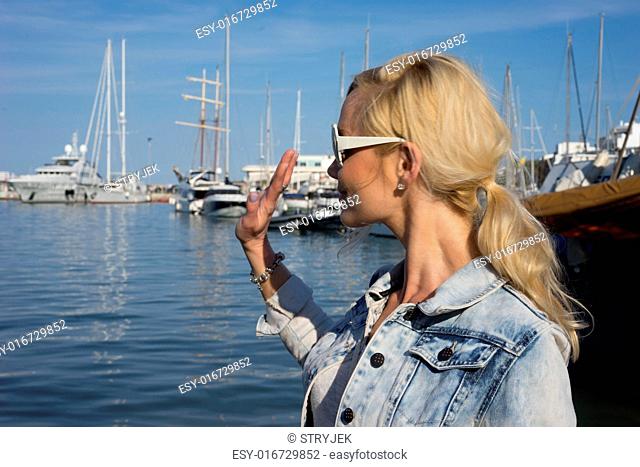 Attractive blond woman wearing sunglasses with her hair in a ponytail standing waving overlooking a harbour full of luxury pleasure boats and yachts