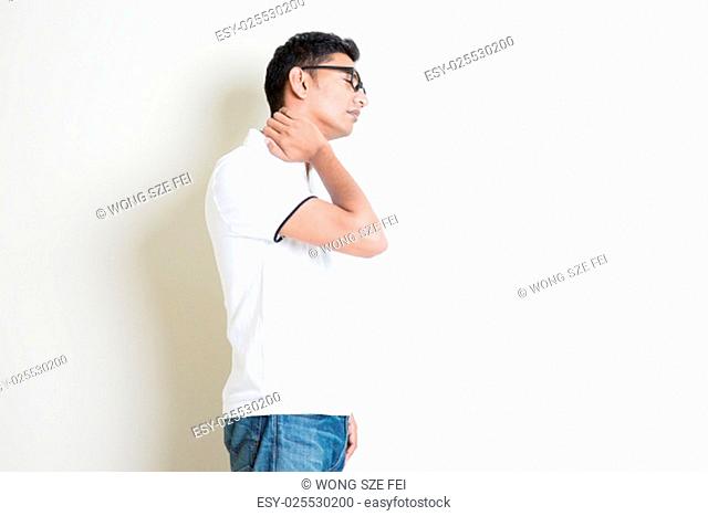 Portrait of Indian guy neck pain, massaging with hand. Asian man standing on plain background with shadow and copy space. Handsome male model