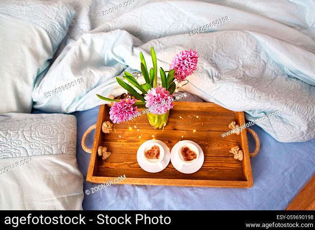 A bouquet of beautiful pink hyacinths is standing in a vase on a tray on which are two cups of cappuccino with a heart pattern, the concept of a surprise in bed