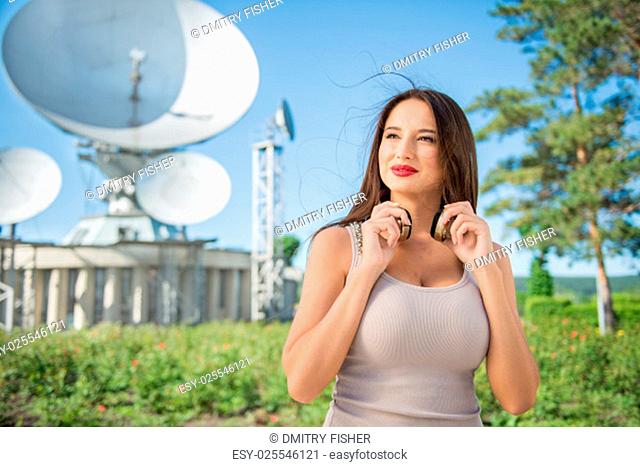 Beautiful young woman wearing vintage music headphones around her neck and standing against background of satellite dish that receives wireless signals from...