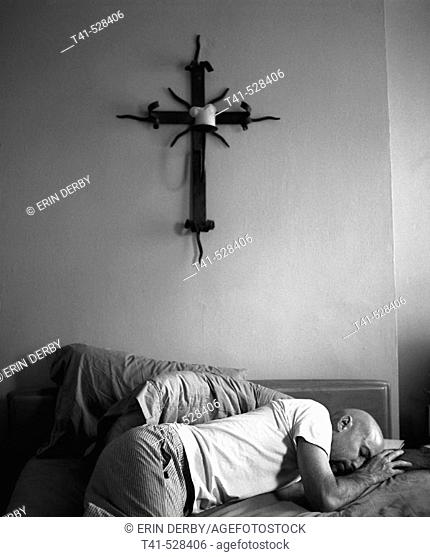 A man in his pajamas on his bed with an iron cross and candle above