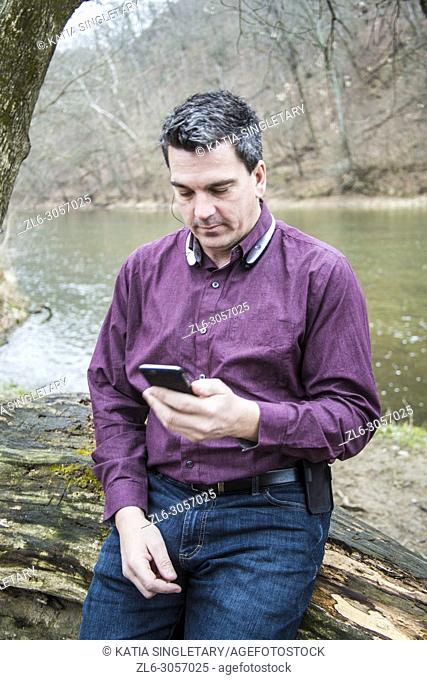 Caucasian handsome dark hair man wearing a purple shirt and his bluetooth. He is in against a wood log in nature and he is focused on his phone