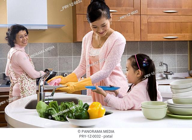 Mother with daughter washing plates while grandmother cooking food in background