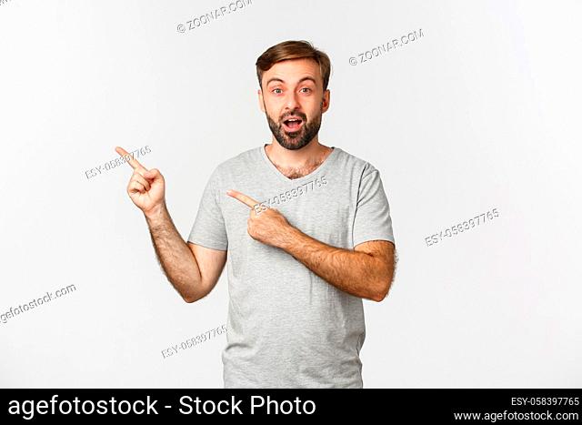 Image of amazed bearded guy in gray t-shirt, pointing fingers at upper left corner logo, showing advertisement, standing over white background