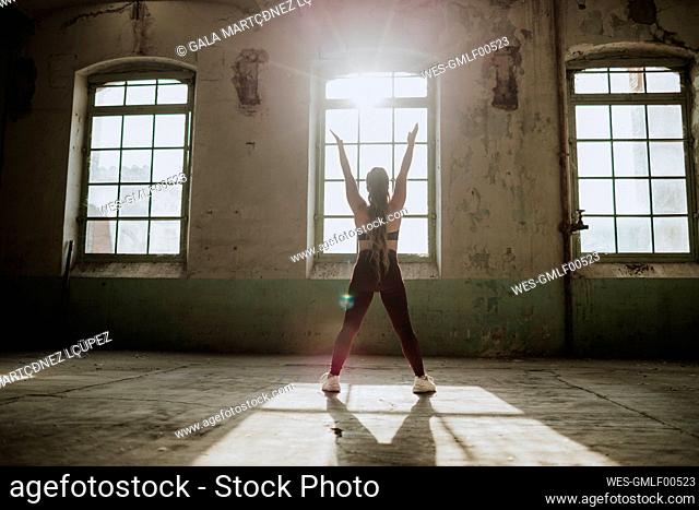 Sportswoman with hand raised exercising against window at abandoned factory