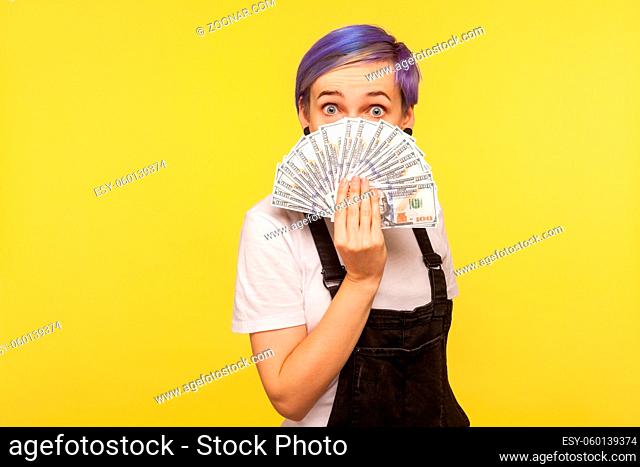 Portrait of fashionable young woman with violet short hair in denim overalls peeking out of money while hiding half face with surprised shocked eyes