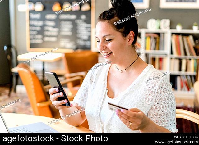 Close-up of smiling young woman holding credit card using smart phone in coffee shop