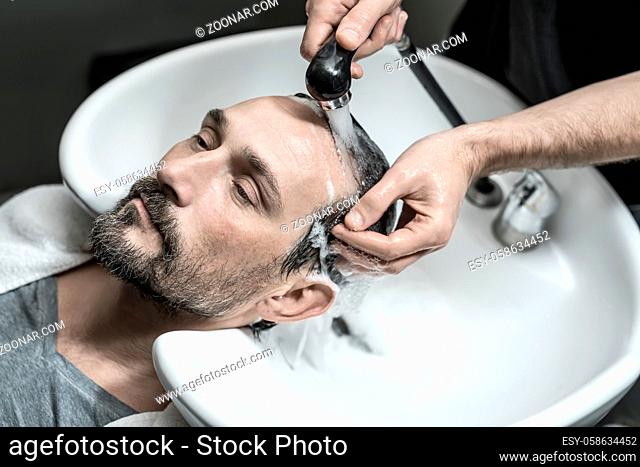 Cute man with a beard lies on the white sink in the barbershop. Barber washes his head with the black faucet. Guy wears a gray T-shirt and a white towel