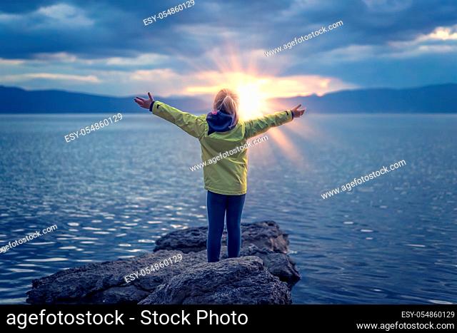 Triumphant little girl lifting her hands high in the air while standing on the large boulders on the shore of Lake Ohrid, Republic of Macedonia