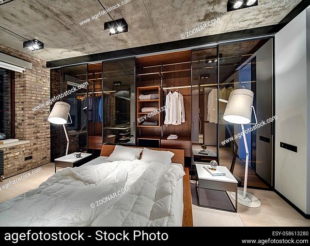 Loft style bedroom with brick wall and concrete ceiling. There is a bed with white pillows and blankets, glowing lamps, wardrobe with glass sliding doors