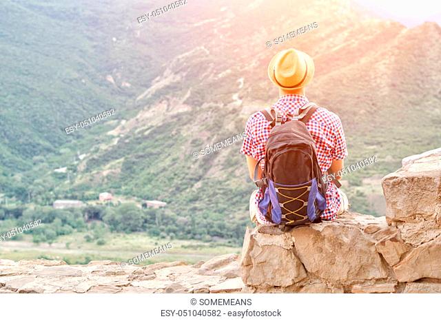 Young man in a hat with a backpack sits on a stone wall against a background of green mountains. Back view