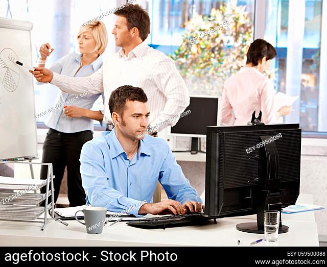 Casual businessman sitting at office desk, working on computer. Businesspeople working in background