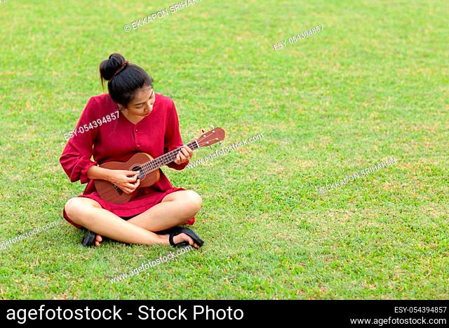 Portrait of beautiful young asian woman with red dress is playing ukulee with feeling happy and smile on green field at the public park