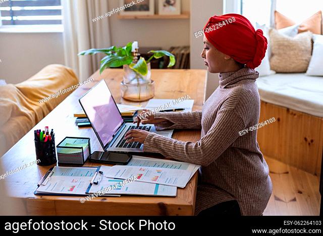 Biracial woman wearing hijab, sitting at table in living room and using laptop with copy space