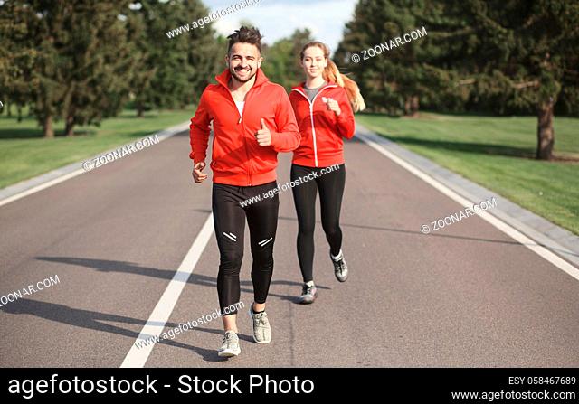 Sport man and woman jogging along road. Beautiful couple in red jackets training and looking at camera. Fitness concept