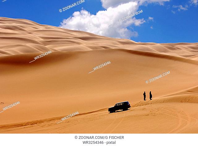 Zwei Personen stehen neben einem Geländefahrzeug in den Weiten der goldgelben Sanddünen im Ubari Sandmeer, Sahara, Libyen / Two personens standing next to their...