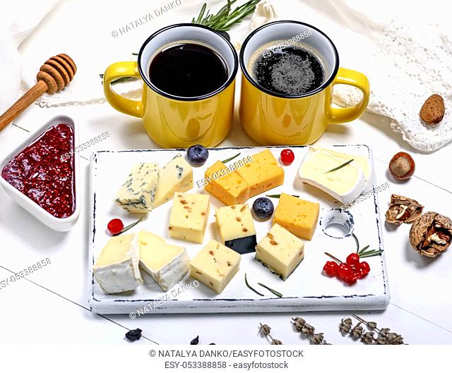 different pieces of cheese on a white wooden board and two yellow cups with a drink, close up